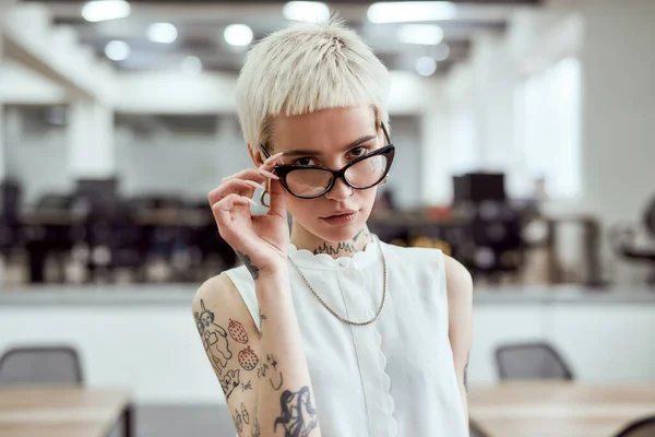 Joven y exitosa. Retrato de mujer de negocios tatuada rubia confiada ajustando sus anteojos y mirando a la cámara mientras está de pie en el espacio de trabajo moderno — Foto de Stock