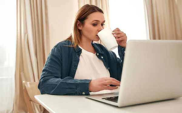 Απομακρυσμένη δουλειά. Νεαρή καυκάσια γυναίκα με casual ρούχα που χρησιμοποιεί φορητό υπολογιστή και πίνει καφέ ή τσάι ενώ κάθεται στο τραπέζι της κουζίνας στο σπίτι, δουλεύοντας εξ αποστάσεως — Φωτογραφία Αρχείου