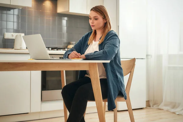 Συγκεντρωμένος στη δουλειά. Νεαρό καυκάσιο κορίτσι σε casual ρούχα που εργάζονται από το σπίτι, κάθεται στο τραπέζι της κουζίνας στο σπίτι και τη χρήση φορητού υπολογιστή — Φωτογραφία Αρχείου