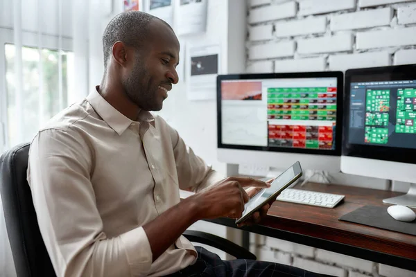 Conoce el mejor futuro aquí. Elegante hombre de negocios africano, comerciante sentado junto al escritorio frente a múltiples monitores y utilizando su teléfono inteligente para comprobar gráficos mientras trabaja en la oficina . —  Fotos de Stock