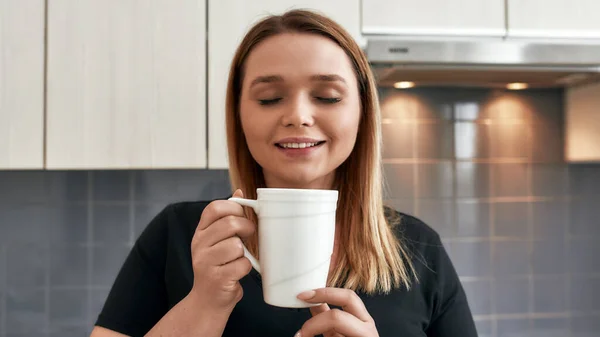 Lieblingsgetränk. Nahaufnahme einer jungen kurvigen Frau, die ihre Augen schließt und lächelt, während sie Tee oder Kaffee in der Küche trinkt — Stockfoto