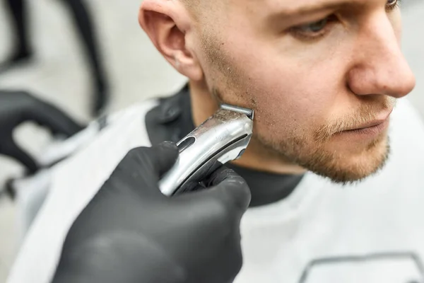 Beard correction. Young handsome man sitting in armchair in barber salon while professional barber trimming beard with electric clipper machine — Stock Photo, Image
