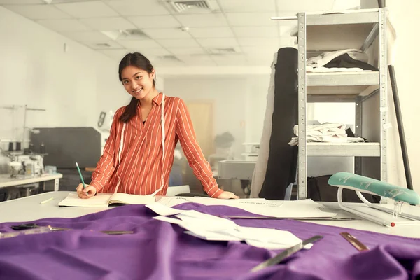 Létrehozod a világod. Fiatal női ruhatervező támaszkodik az asztalra szövet, textil és varrás kellékek rajta, miközben tervezi az új gyűjtemény és írás jegyzetek — Stock Fotó