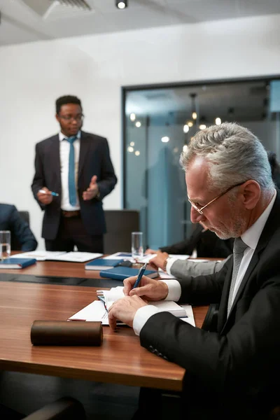 Focado maduro caucasiano empresário escrevendo algo em seu bloco de notas, tendo uma reunião no escritório moderno — Fotografia de Stock