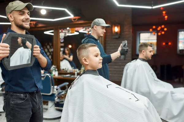 Coupe de cheveux pour petit garçon. Jeune coiffeur souriant tenant un miroir et montrant le résultat de son travail à mignon petit garçon assis dans la chaise de salon de coiffeur. Enfant visitant salon de coiffure — Photo