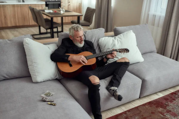 Laat de muziek stromen. Baard man van middelbare leeftijd, artiest die liedjes schrijft, gitaar speelt, op de bank zit. Metalen pijp voor het roken van marihuana, cannabisknoppen in een plastic zak en aansteker liggend op de bank — Stockfoto