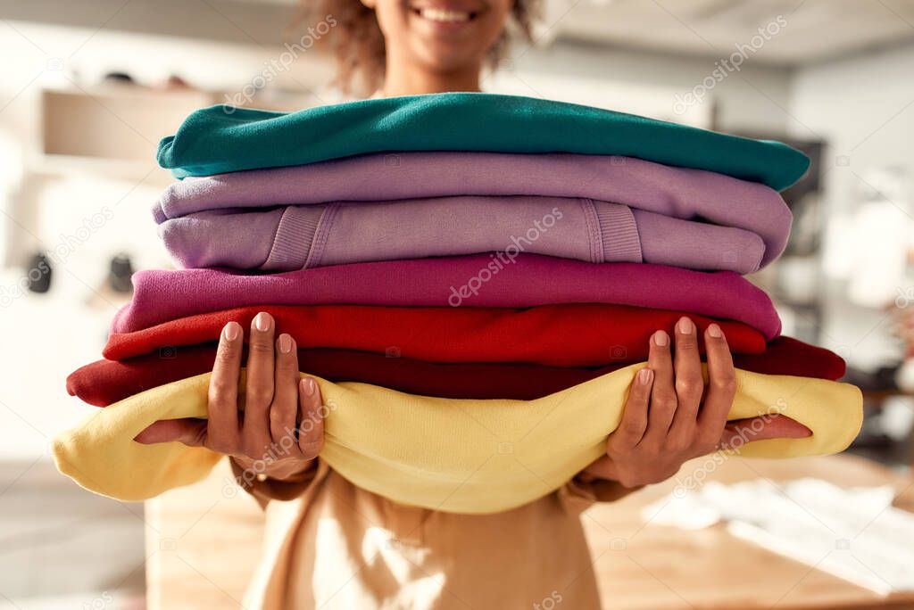 Cropped shot of woman holding stack of colorful sweatshirts, hoodies while standing in the store. Clothes at custom T-shirt, clothing printing company. Horizontal shot