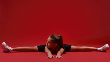 Esneme süresi. Şirin bir çocuk, kız sporla meşgul, jimnastikçi de bacaklarını ayırırken yana bakıyor. Kırmızı arka planda izole edilmiş. Fitness, eğitim, aktif yaşam tarzı konsepti. Yatay çekim