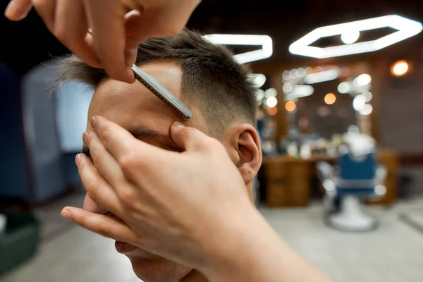 Erkek tıraşı. Berber ellerin düz jilet tutarken ve genç beyaz adam için modern saç kesimi yaparken yakın çekilen bir fotoğraf. Berber dükkanında saçını kestiriyorum. — Stok fotoğraf