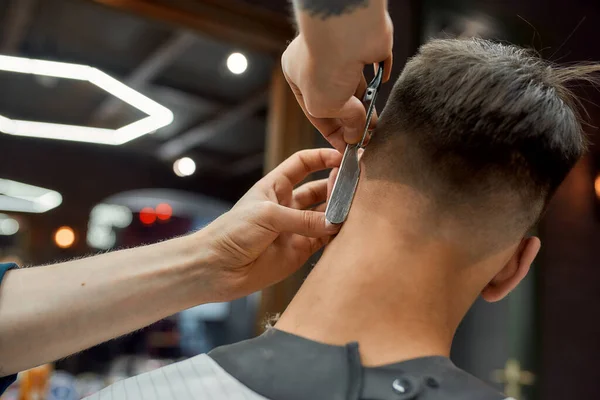 Високоякісна робота. Барбер тримає пряму бритву і робить сучасну зачіску для свого клієнта, працюючи в перукарні. — стокове фото