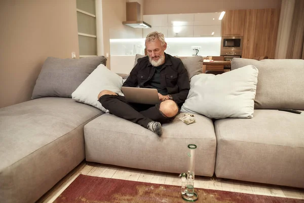 Binnen aan het werk. Een man van middelbare leeftijd die laptop gebruikt terwijl hij op de bank zit. Bong of glazen waterpijp, knoppen in plastic zak en aansteker in zijn buurt. Cannabis en wiet legalisatie concept — Stockfoto