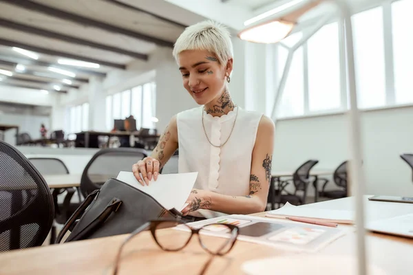 Megtaláltam. Fiatal boldog szőke tetovált üzletasszony rövid frizurával kivesz néhány iratot a táskájából és mosolyog, miközben az íróasztalánál ül az irodában. — Stock Fotó