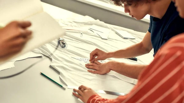Production process. Male hands working with sketches or patterns at studio and preparing fabric. Group of millennials creating clothes together — Stock Photo, Image