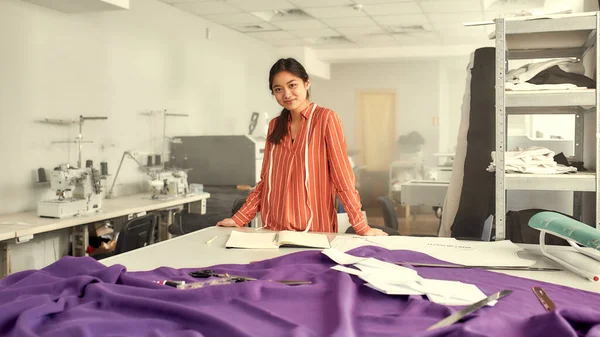 Meg tudom csinálni. Fiatal női ruhatervező támaszkodik az asztalra szövet, textil és varrás kellékek rajta, miközben tervezi az új gyűjtemény és írás jegyzetek — Stock Fotó