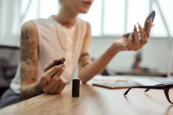 Τελειότητα. Πορτρέτο της νεαρής και κομψής ξανθιάς επιχειρηματία με τατουάζ προσαρμόζοντας το μακιγιάζ της, εφαρμόζοντας κραγιόν ενώ κάθεται στο σύγχρονο γραφείο — Φωτογραφία Αρχείου