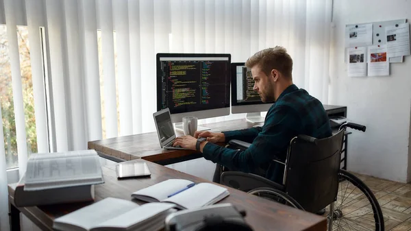 IT professional. Young caucasian male web developer in a wheelchair writing program code while sitting at workplace with three monitors in the modern office. HTML, PHP and javascript — Stock Photo, Image