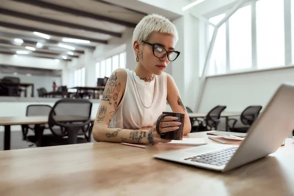 Az üzleten gondolkodom. Fiatal gondolkodó tetovált üzletasszony szemüveg kezében egy csésze és nézi laptop képernyőn, miközben ül egyedül a modern irodában. — Stock Fotó