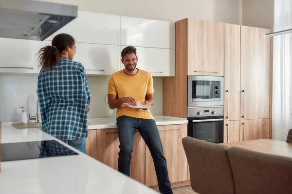 On adore notre maison. Heureux couple multiculturel en vêtements décontractés essuyant les plats et parlant entre eux tout en se tenant dans leur cuisine moderne. Nettoyage maison — Photo