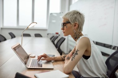 İş kadını işine odaklanmış. Kısa saçlı, genç ve güzel bir kadının toplantı odasında çalışırken laptopunu kullanmasının yan görüntüsü.