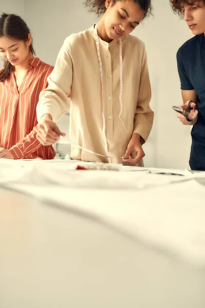 Consigue lo mejor. Joven diseñadora de moda femenina alegre midiendo tela, material textil antes de cortarlo en un estudio. Grupo de millennials creativos trabajando juntos — Foto de Stock