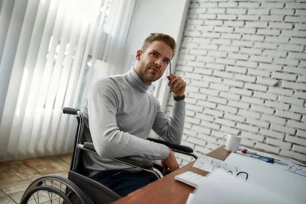 Wachten op inspiratie. Portret van een jonge serieuze mannelijke architect in een rolstoel die ergens aan denkt terwijl hij met blauwdruk werkt op zijn werkplek in het moderne heldere kantoor — Stockfoto