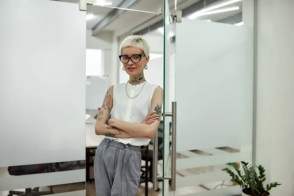 Before meeting. Young, happy beautiful blonde tattooed businesswoman leaning on the glass door and looking at camera with smile while standing in modern office