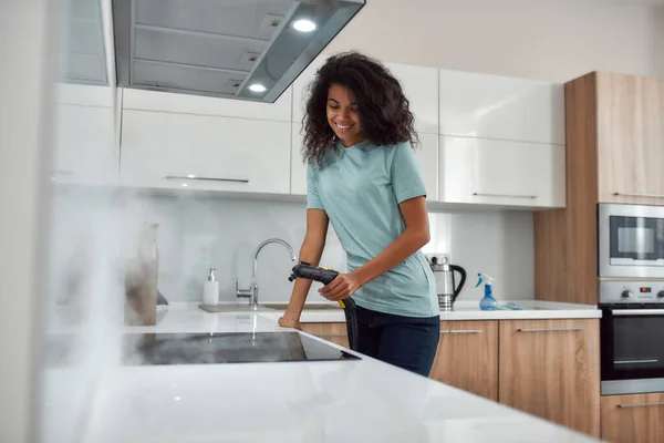 Servizio professionale. Ritratto di una giovane donna afro americana in uniforme pulizia stufa elettrica con pulitore a vapore e sorridente mentre lavora nella moderna cucina — Foto Stock