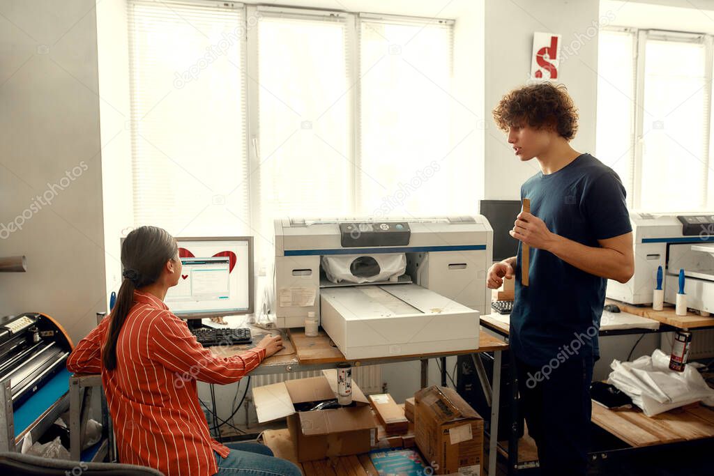 Because we love designing. Young workers, man and woman putting t-shirt for printing in the silk screen printing machine at workplace. Woman sending chosen file to printing machine