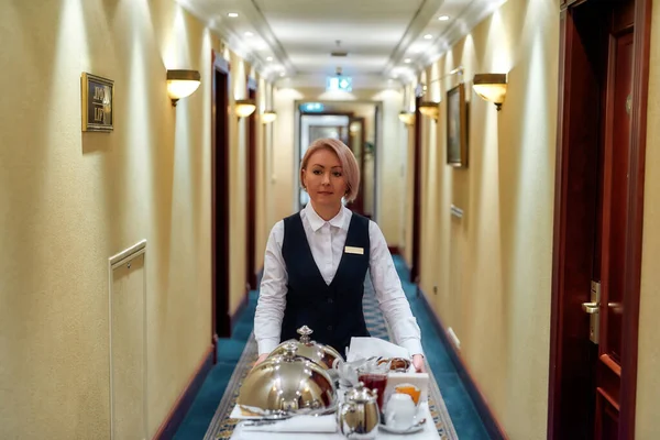 Experimente a paixão da hospitalidade. Garçonete de uniforme andando ao longo do corredor enquanto entrega bandeja com comida em um quarto de hotel. Serviço de quarto . — Fotografia de Stock
