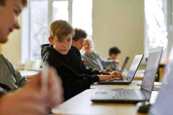 Αναζητώ βοήθεια. Πορτρέτο του καυκάσιου σχολιαρόπαιδου που κοιτάζει το φορητό υπολογιστή του γείτονά του ενώ κάθεται σε μια τάξη με άλλους μαθητές κατά τη διάρκεια ενός μαθήματος στο σύγχρονο έξυπνο σχολείο — Φωτογραφία Αρχείου