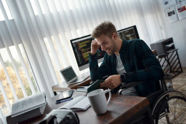 Boekenliefhebber. Portret van een vrolijke mannelijke webontwikkelaar in een rolstoel die iets leest en glimlacht terwijl hij op zijn werkplek zit met meerdere computerschermen op de achtergrond — Stockfoto