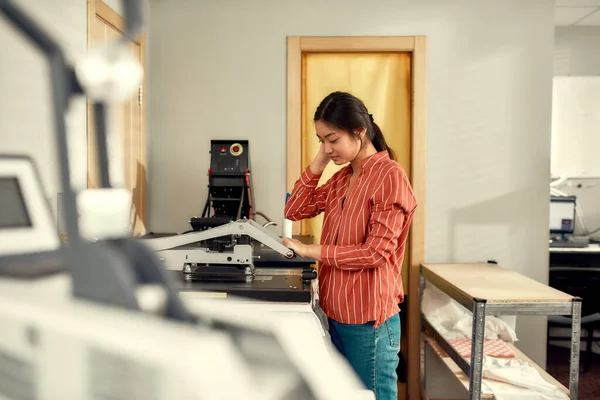 Quality printing. Young cheerful female worker using heat press transfer machine for printing advertisement sticker with text on t-shirt at workplace