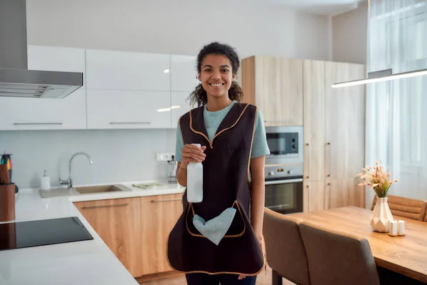 Profesjonalny serwis. Młoda, wesoła afro amerykańska kobieta trzymająca spray detergent, patrząca w kamerę i uśmiechnięta podczas sprzątania w kuchni. Profesjonalna sprzątaczka w mundurze — Zdjęcie stockowe