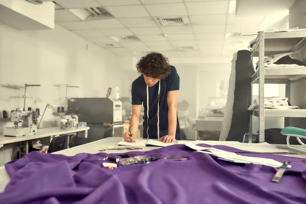 Powiększ widok. Młody projektant odzieży męskiej opierający się o biurko z materiałami, materiałami włókienniczymi i wyrobami do szycia, planując jednocześnie nową kolekcję i notatki do pisania — Zdjęcie stockowe