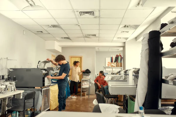 Not delivering, making. Full-length shot of young cheerful millennials using heat press transfer, silk screen printing machine for printing t-shirts at workplace