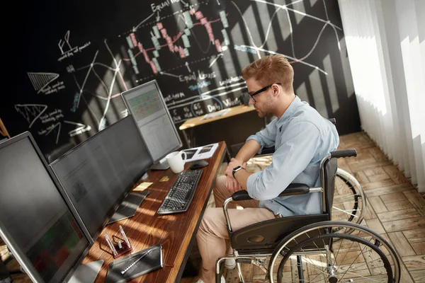 Ik werk de hele dag. Zijaanzicht van jonge succesvolle mannelijke handelaar in een rolstoel, kijkend naar grafieken en financiële rapporten op meerdere computerschermen tijdens zijn werk op zijn werkplek in het moderne kantoor — Stockfoto