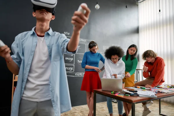 Potente herramienta para los negocios. Joven hombre asiático en ropa casual vistiendo auriculares de realidad virtual mientras sus colegas discuten nuevo proyecto detrás de él. Alta tecnología —  Fotos de Stock