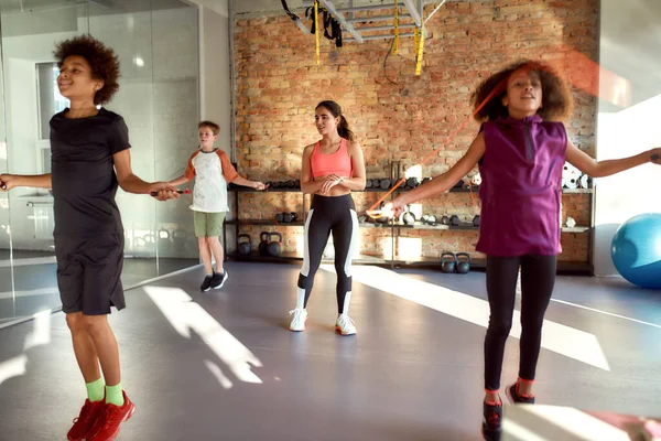 Energiza tu vida hoy. Tirador de cuerpo entero de entrenadora controlando, cuidando a los niños, mientras están saltando la cuerda en el gimnasio. Deporte, vida sana, concepto de educación física —  Fotos de Stock