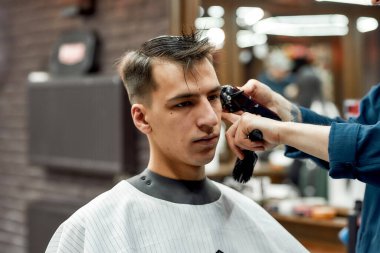 Berber dükkanında. Genç yakışıklı adam berber dükkanında saçını kestiriyor. Berber saç makasıyla çalışıyor.