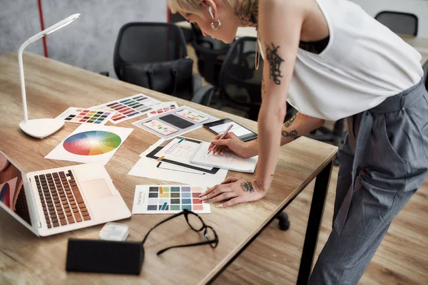 Mujer creativa. Joven rubia tatuada diseñadora femenina haciendo algunos bocetos mientras está de pie cerca del escritorio de la oficina. Trabajar con muestras de muestras de muestras de color — Foto de Stock