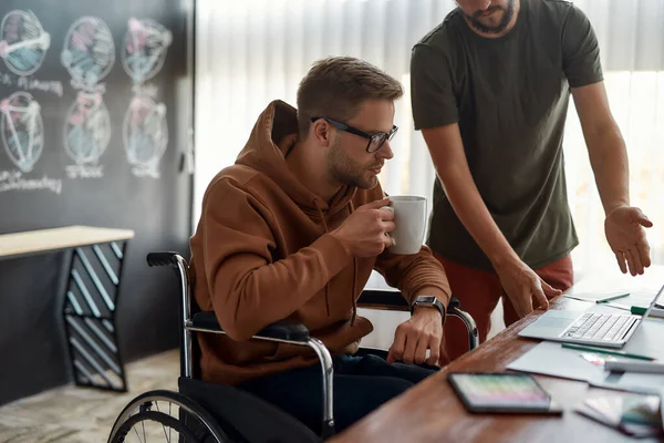 新しいプロジェクトに取り組む。若い男性デザイナーが車椅子でコーヒーを飲みながら、クリエイティブなオフィスで一緒に仕事をしながら同僚といくつかのアイデアを話し合う — ストック写真