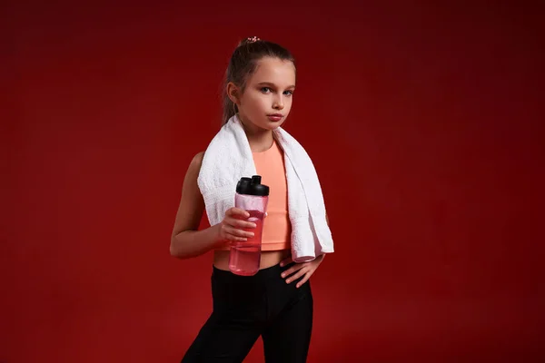 Bebe más. Una linda chica dedicada al deporte, mirando a la cámara mientras está de pie con una toalla y una botella de agua. Aislado sobre fondo rojo. Fitness, entrenamiento, concepto de estilo de vida activo. Disparo horizontal — Foto de Stock