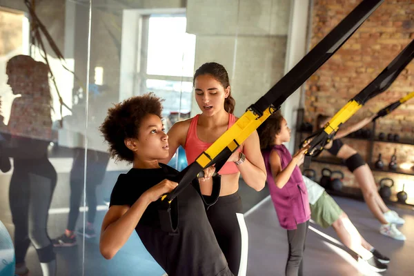 Continuă. Portretul antrenorului de sex feminin care ajută adolescentul în timp ce se antrenează folosind curele de fitness în sala de sport. Sport, stil de viață sănătos, concept de educație fizică — Fotografie, imagine de stoc