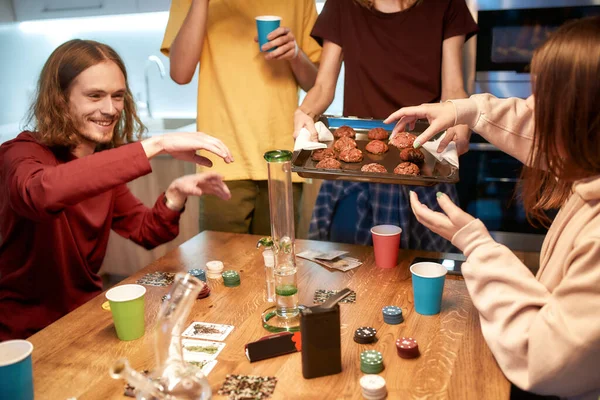 Hacer galletas de marihuana. Jóvenes felices comiendo galletas con cannabis mientras están sentados en la cocina en casa y jugando al póquer. Herramientas de marihuana sobre la mesa — Foto de Stock