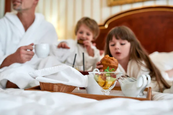 Saludable y sabroso. Familia con albornoces blancos desayunando en la cama, comiendo pasteles y tomando café en la lujosa habitación del hotel. Concéntrate en la taza de fruta en la bandeja. Comida, resort, concepto de servicio de habitaciones — Foto de Stock