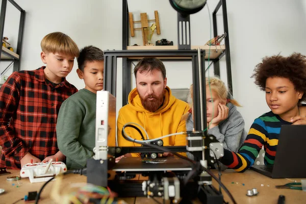 Experimente la innovación digital. Grupo de niños inteligentes escuchando a su profesor masculino mientras mira la impresora 3D y el prototipo de detalle de plástico en la lección de la escuela de robótica — Foto de Stock