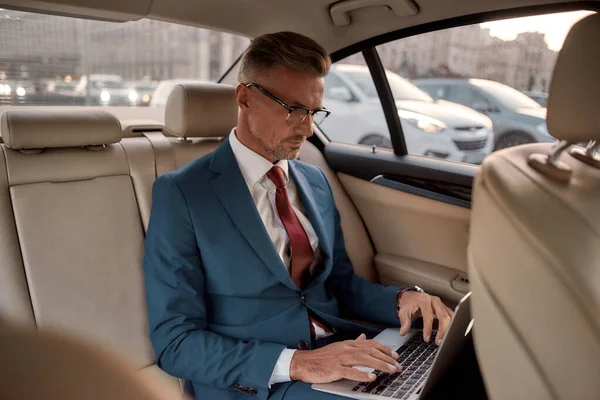 Doelen stellen. Ernstige volwassen zakenman in volle pak werken op zijn laptop terwijl zitten in de auto — Stockfoto