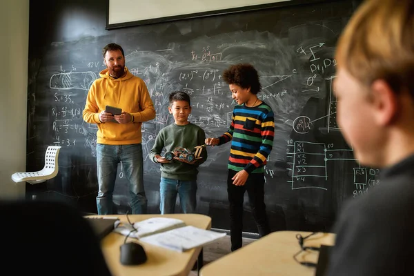 Una escuela de calidad. Joven profesor de ciencias de pie cerca de la pizarra, sosteniendo la tableta PC y mirando a sus estudiantes mientras están demostrando su propio vehículo robot — Foto de Stock