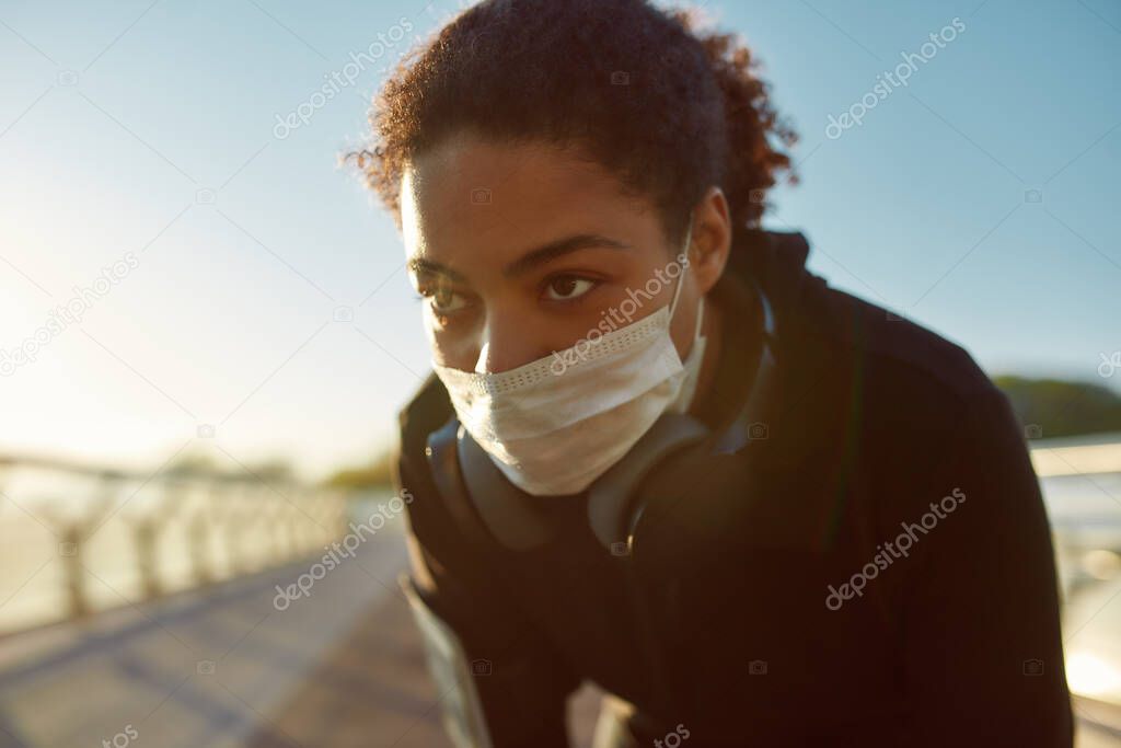 Close up photo of exhausted african female jogger in medical mask resting after run while standing on the bridge in the morning