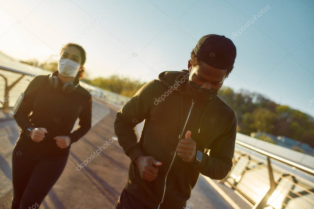 Staying fit. Young african athletic family in medical masks running on the bridge during a pandemic. Sport and coronavirus. Covid-19